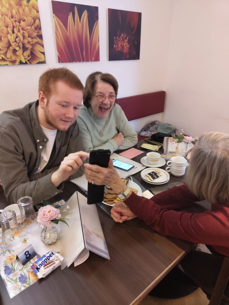 Ehrenamtlicher erklärt zwei Senior*innen am gedeckten Kaffeetisch den Umgang mit einer Smartphone-App
