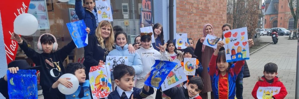 Gruppenfoto mit Rim Alabdallah und dem Basteltreff vor dem Café Credo. Kinder halten selbstgemalte Bilder in die Höhe.
